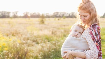 Little baby boy and his mother walking in the fields during summer day. Mother is holding and tickling her baby, babywearing in sling. Natural parenting concept